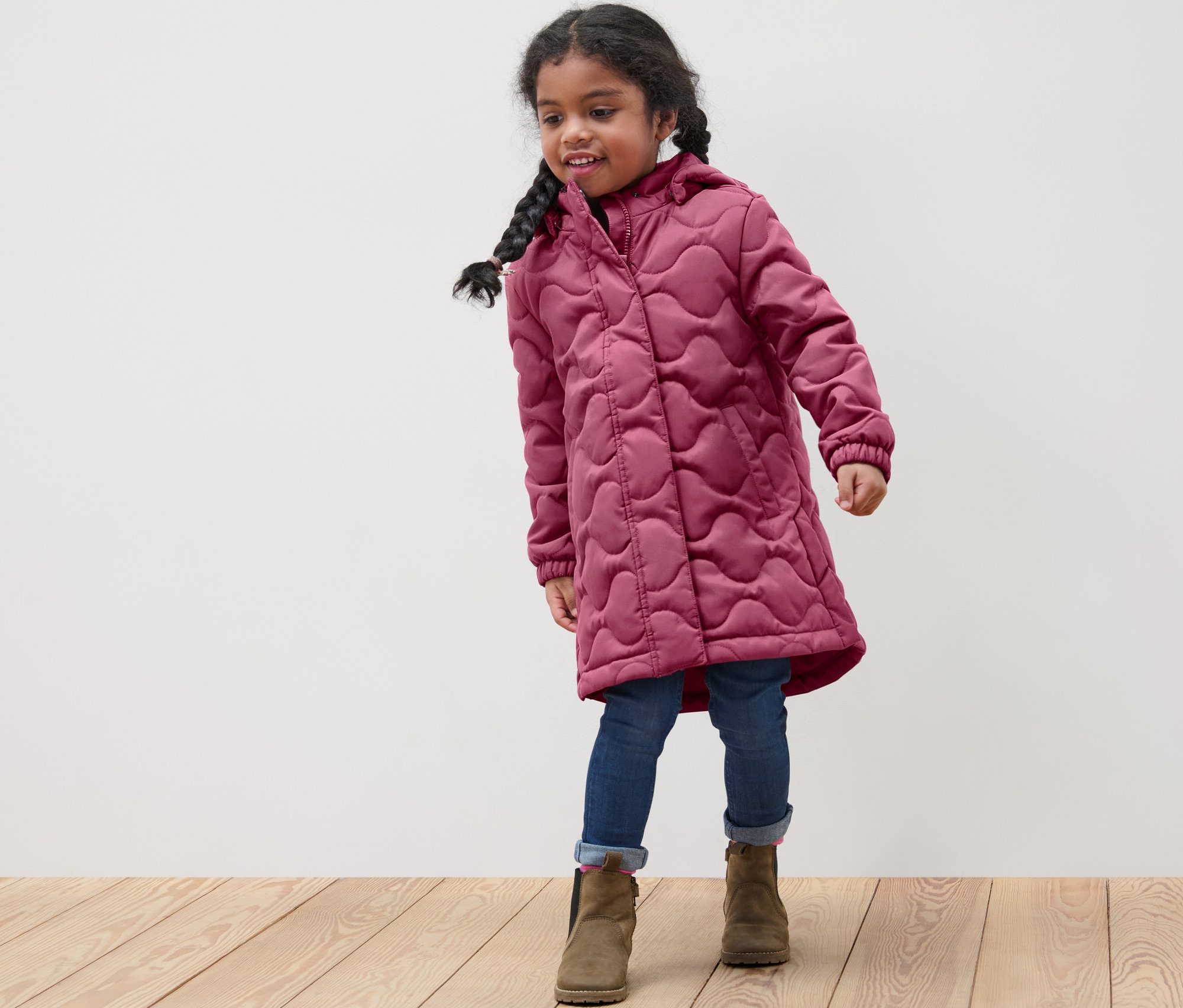 Mädchen trägt einen Kinder-Steppmantel, Jeans und Stiefel.