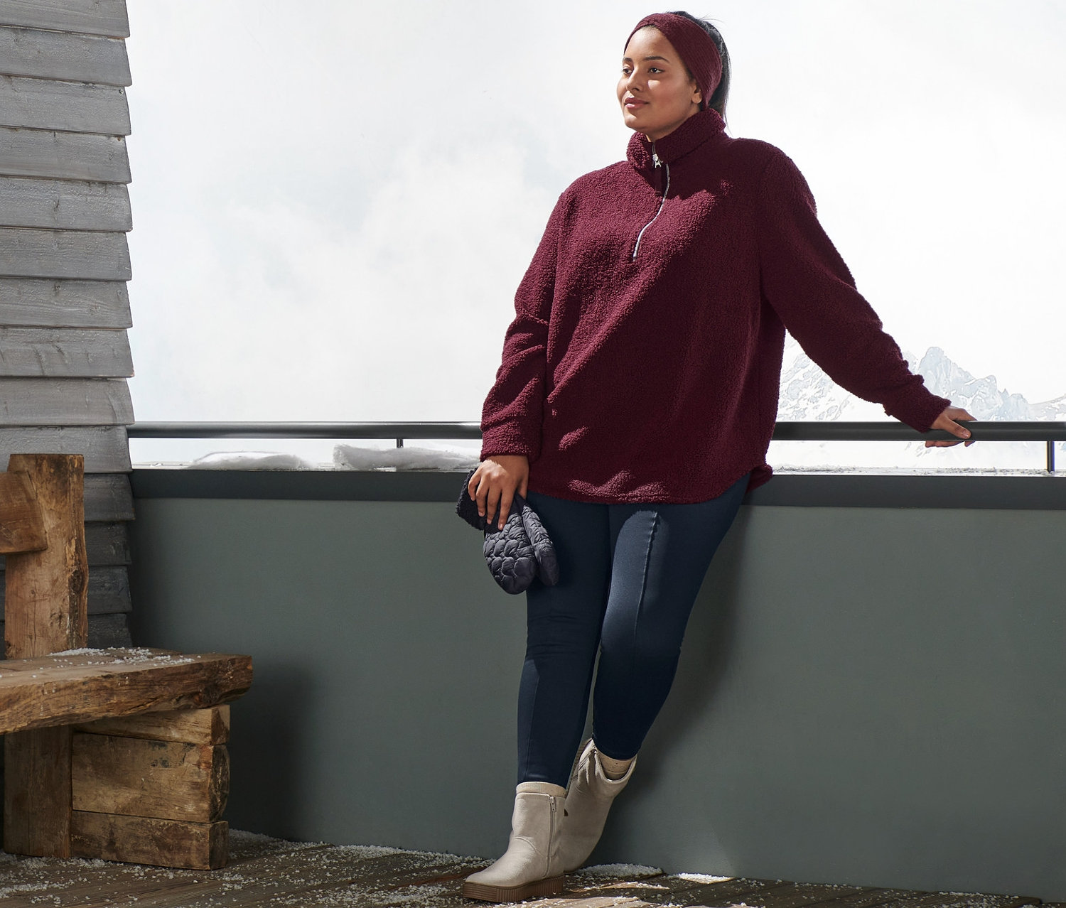 Eine Frau lehnt an einem Geländer und trägt einen Oversized Teddypullover, ein bordeauxrotes Strick-Stirnband, blaue Thermotights und Veloursleder-Boots. Sie hält Stepp-Fäustlinge.
