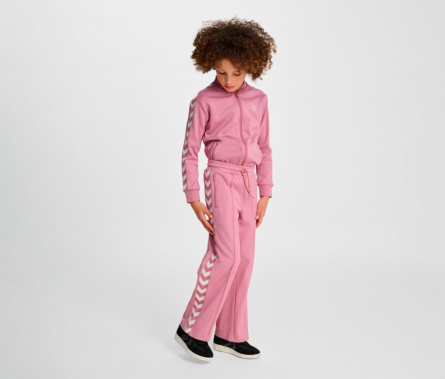Mädchen in rosa Hummel HMLJR Archive Wideleg Poly Pants Anzug steht vor hellem Hintergrund.
