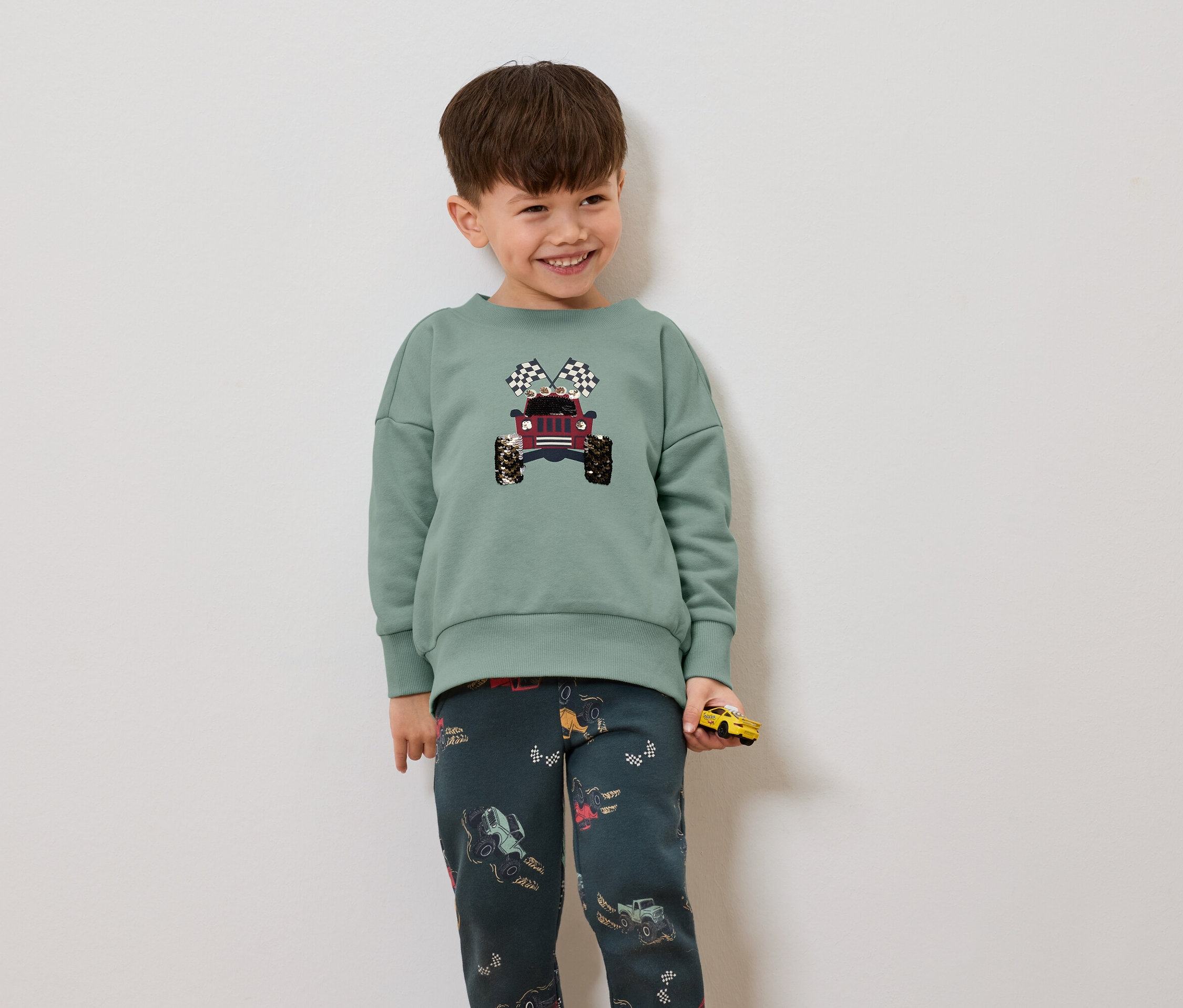 Junge trägt einen grünen Pullover mit Jeep- und Zielflaggendruck, dazu dunkle Hosen mit Jeep-Muster und hält ein gelbes Spielzeugauto.