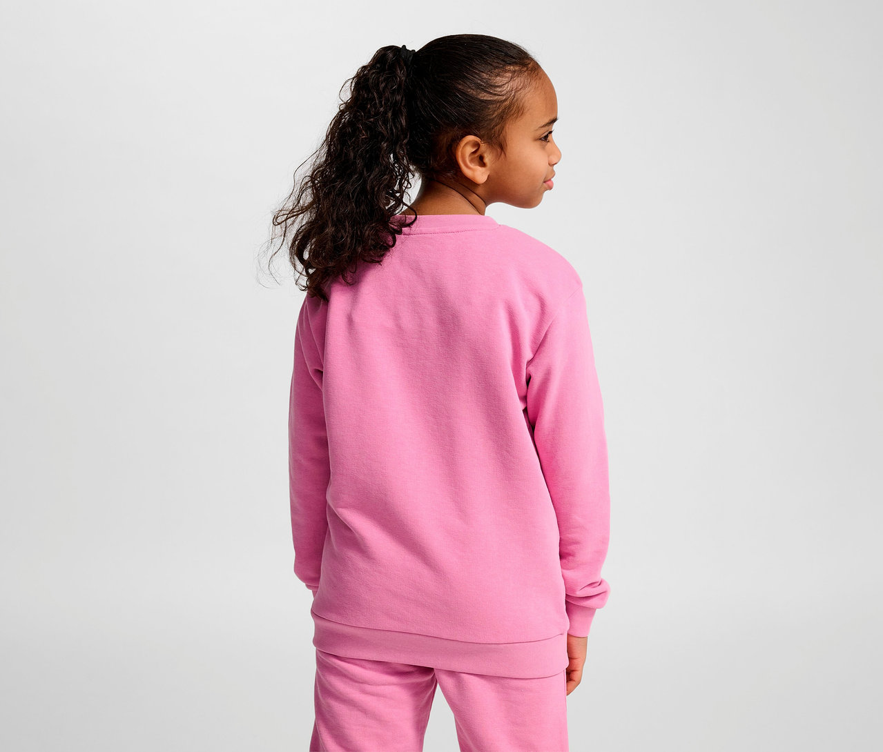 Mädchen mit Pferdeschwanz posiert in einem rosa Trainingsanzug. Sie trägt ein rosa HUMMEL HMLJR Base-Sweatshirt.
