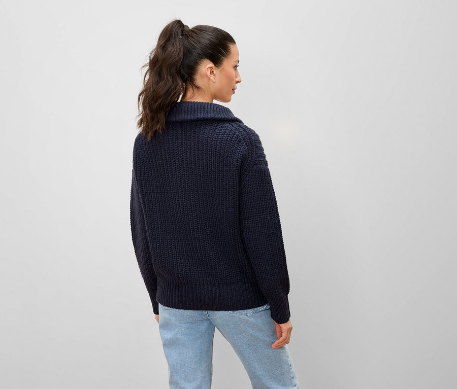 Frau mit Pferdeschwanz steht mit dem Rücken zur Kamera und trägt einen blauen Strickpullover und Jeans.