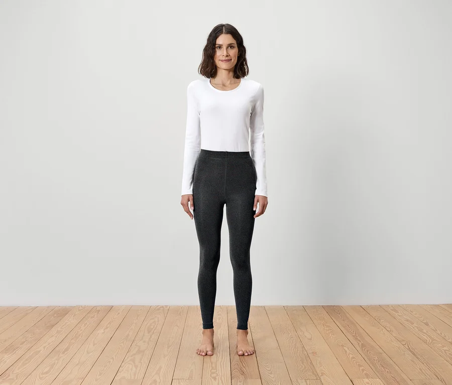 Frau steht barfuß auf Holzboden und trägt ein weißes Langarmshirt und graue Leggings.