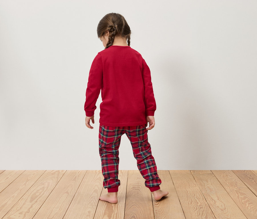 Mädchen mit Zöpfen trägt einen roten, langärmeligen Schlafanzug und karierte Hosen, steht mit dem Rücken zum Betrachter auf einem Holzboden.