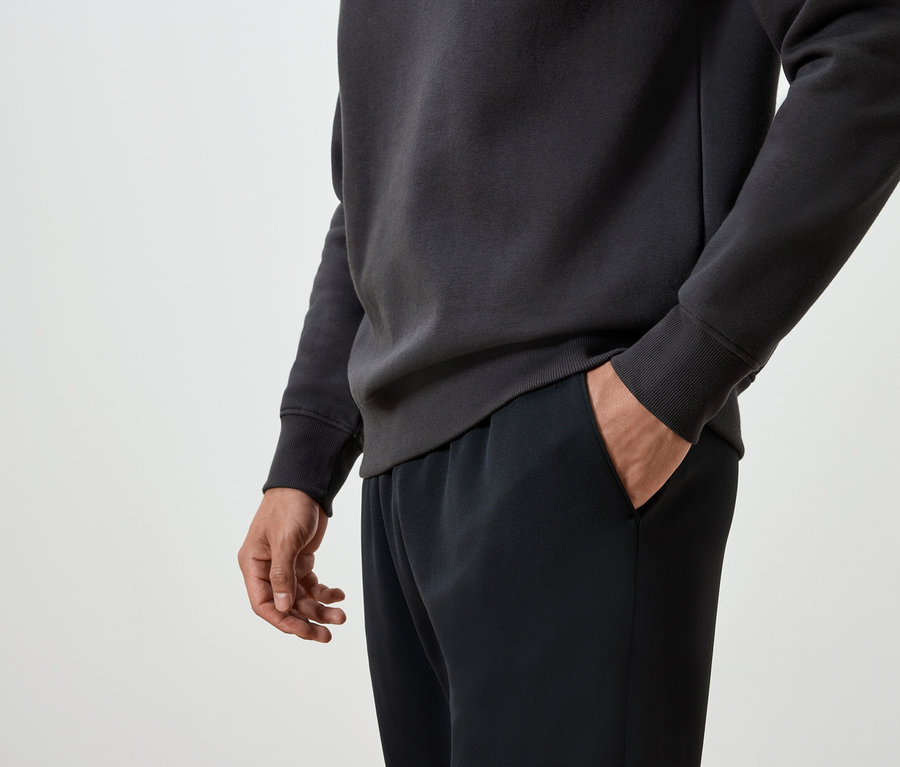 Mann hat Hand in der Tasche einer schwarzen Trainingshose und trägt einen dunkelgrauen Pullover.