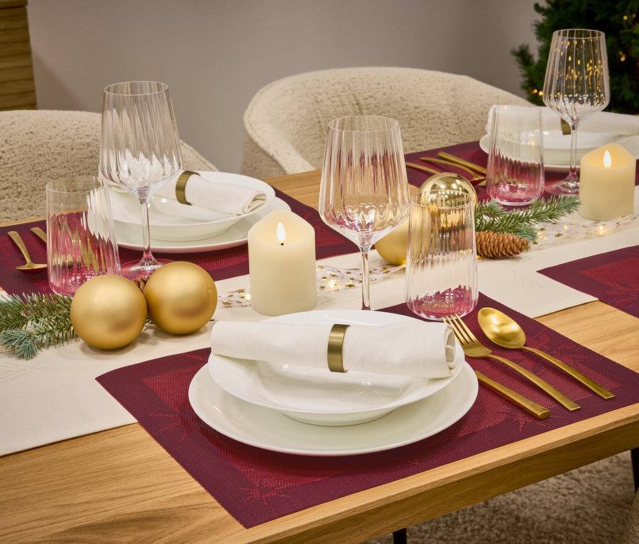 Ein gedeckter Tisch mit weinroten Platzsets, weißem Geschirr, Gläsern, goldenem Besteck, Kerzen und Dekorationen.