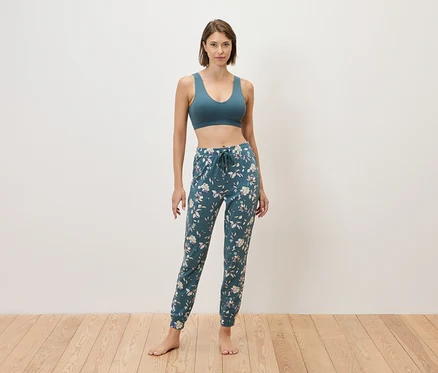 Frau steht barfuß vor einer Wand und trägt einen blauen Sport-BH und eine blaue Jogginghose mit Blumenmuster.
