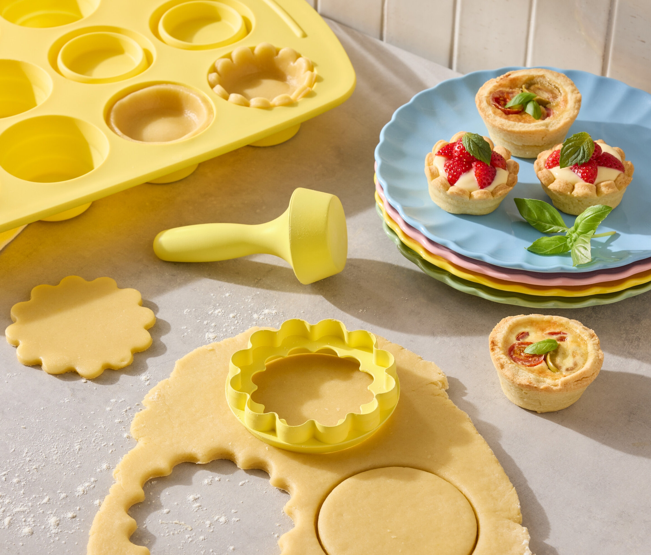 Gelbe Tarteform mit Ausstechern und fertige Tartelettes auf bunten Tellern.