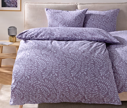 Bettwäsche mit lila Blättermotiv auf einem Bett mit beigem Kopfteil.