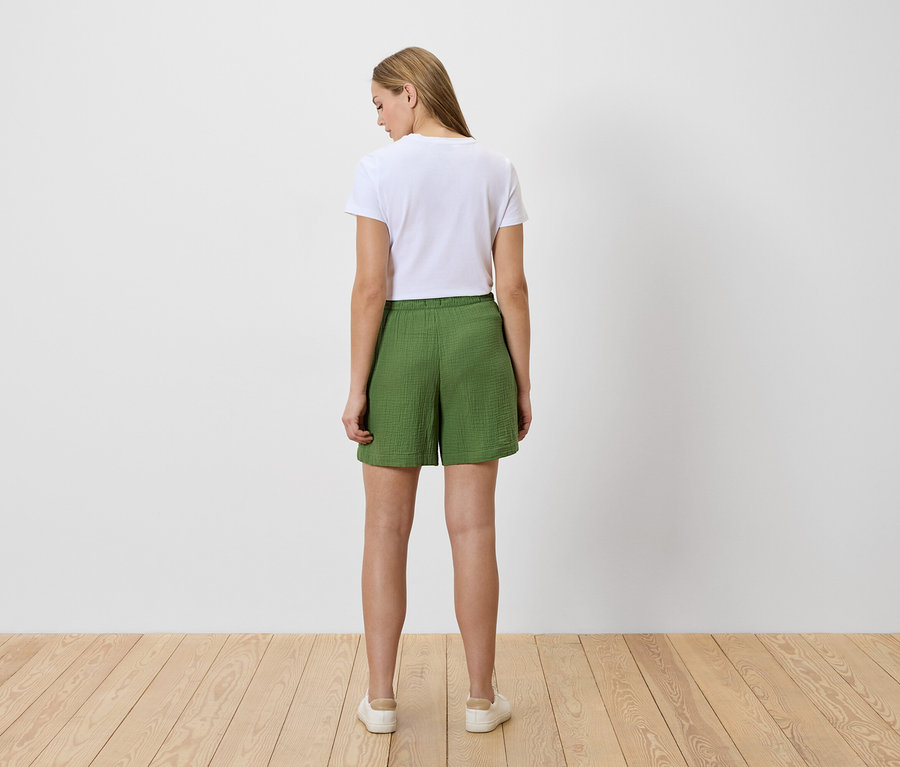 Rückansicht einer Frau mit weißem T-Shirt und grünen Shorts, die auf einem Holzboden steht.