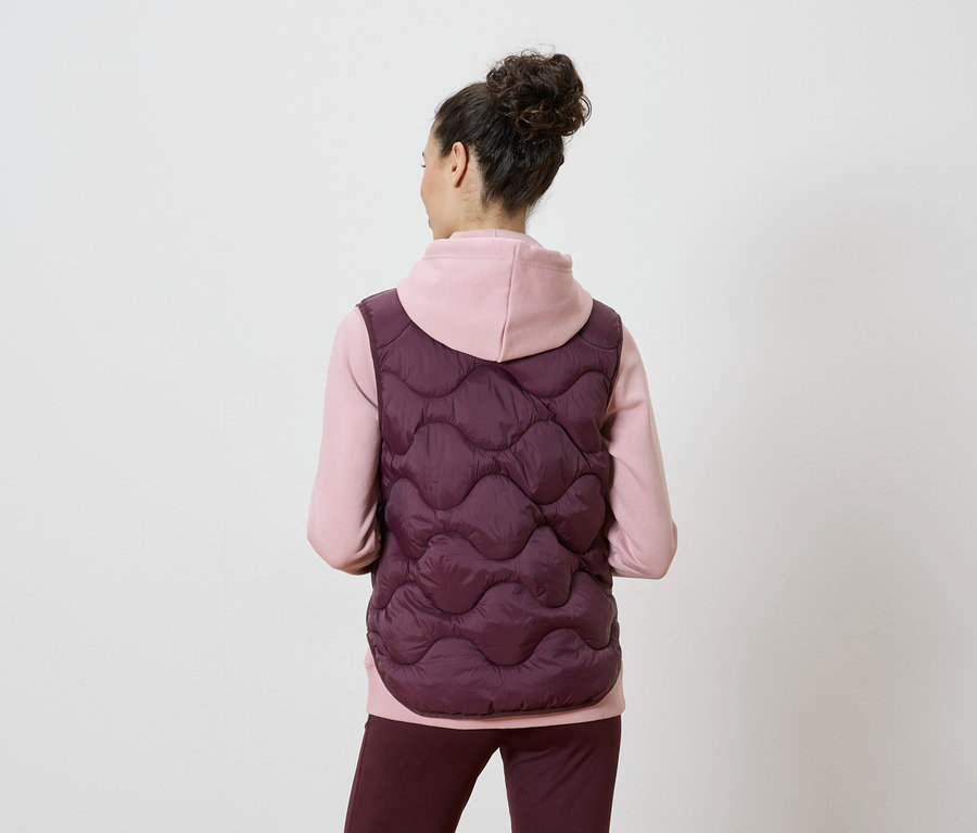Rückansicht einer Frau mit Dutt, die eine bordeauxfarbene Steppweste über einem rosa Hoodie und einer bordeauxfarbenen Hose trägt.