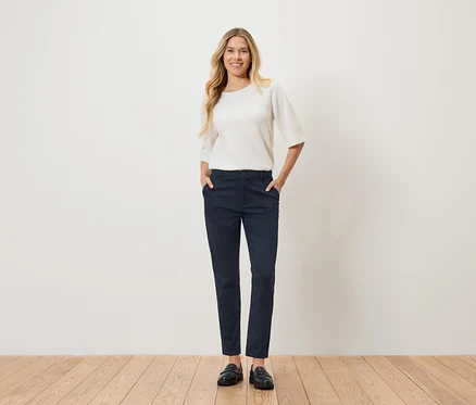 Eine Frau mit blonden, gewellten Haaren steht mit den Händen in den Hosentaschen da. Sie trägt einen weißen Pullover, eine marineblaue Hose und schwarze Schuhe.