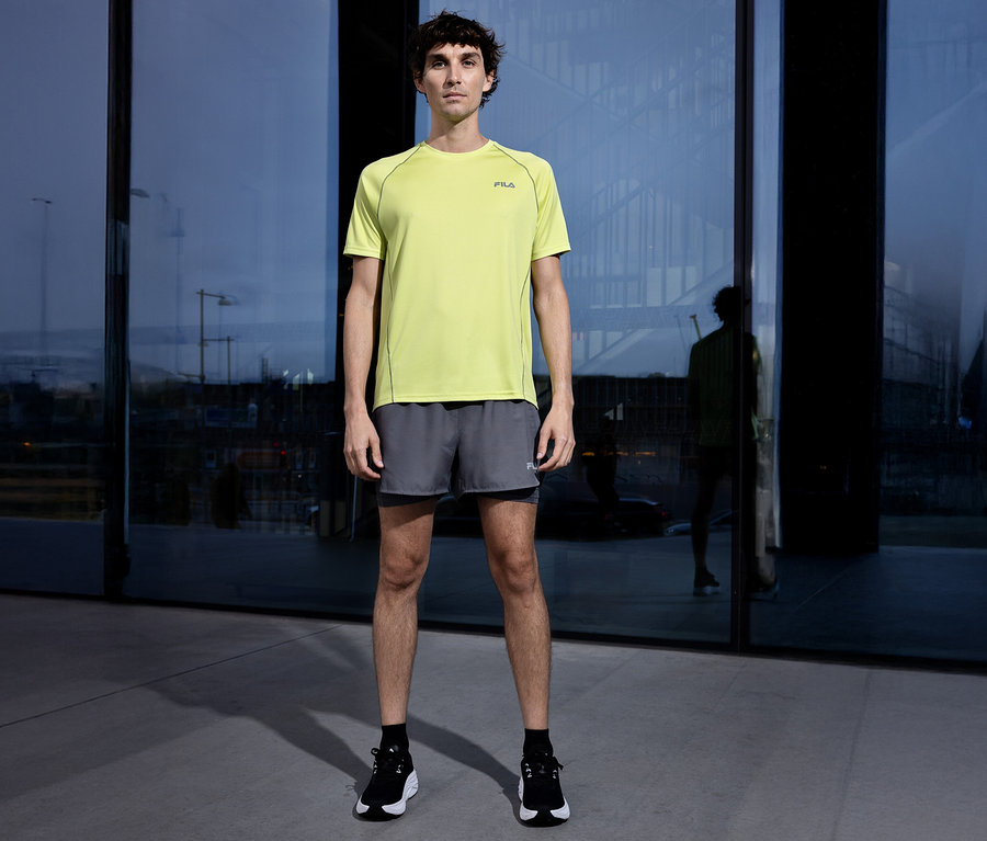 Mann steht vor einem Gebäude und trägt ein gelbes Fila-T-Shirt, graue Shorts und schwarze Schuhe.
