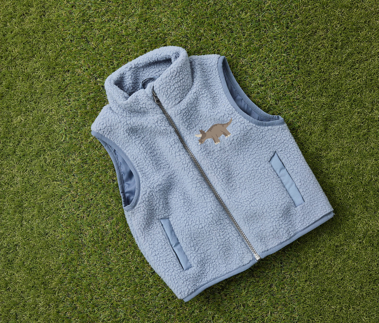 Die hellblaue NAME IT Kinder-Teddyweste mit Reißverschluss und Dino-Motiv liegt auf grünem Gras.