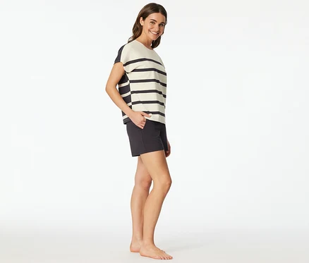 Frau trägt ein SCHIESSER Shorty-Set mit gestreiftem Oberteil und dunklen Shorts, lächelt und hat die Hand in der Tasche.