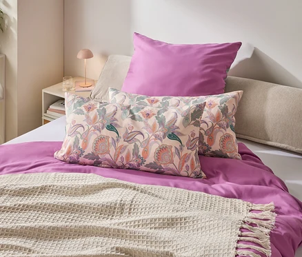 Ein Schlafzimmer mit einem Bett, das mit einem lila Bettbezug, Kissen mit Blumenmuster und einer cremefarbenen Decke mit Fransen geschmückt ist.