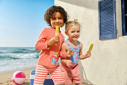 Zwei Kinder mit Eis am Strand. Das Mädchen trägt einen Kinder-Badeanzug mit Seesternen und Jeansshorts. Der Junge trägt eine Kinder-Kombi mit UV-Schutz und Krake.