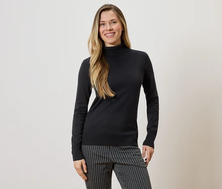 Frau mit langen blonden Haaren, die einen schwarzen Rollkragenpullover und eine gestreifte Hose vor einem hellen Hintergrund trägt.