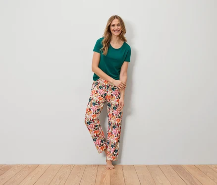 Eine Frau steht barfuß vor einer Wand. Sie trägt ein grünes T-Shirt und eine farbenfrohe Hose mit Blattmuster.