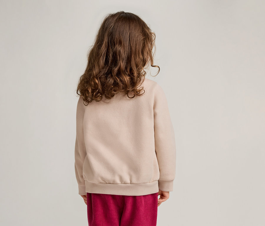 Rückansicht eines Kindes mit lockigem Haar, das einen beigen Pullover und eine rote Cordhose trägt.