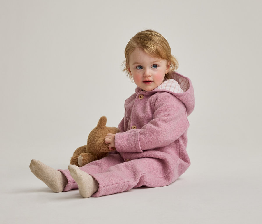 Ein blondes Kleinkind mit blauen Augen sitzt mit einem Teddybären in einem rosa Overall mit Kapuze auf dem Boden.
