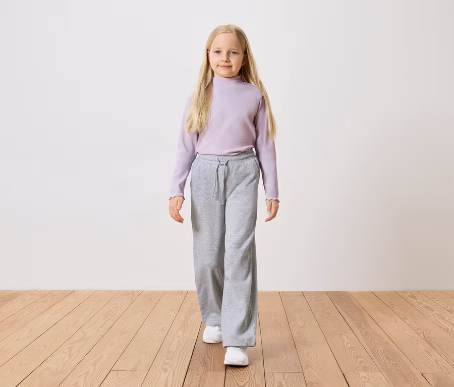 Mädchen mit langen blonden Haaren, trägt ein lila Langarmshirt, graue weite Hosen und weiße Turnschuhe.