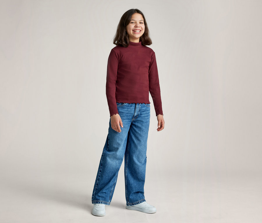 Ein junges Mädchen mit dunklen Haaren lächelt, trägt ein weinrotes Langarmshirt mit Stehkragen, blaue weite Jeans und weiße Turnschuhe vor hellem Hintergrund.