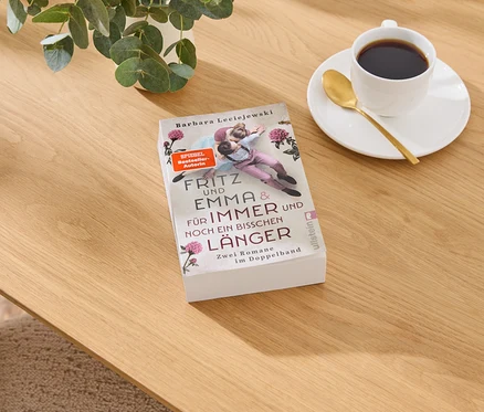 Buch „Fritz und Emma & Für Immer und Noch Ein Bisschen Länger“ von Barbara Leciejewski auf Holztisch, daneben Kaffeetasse und Pflanze.