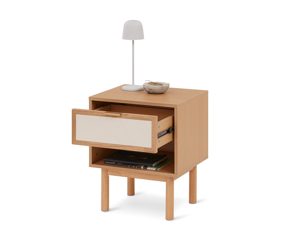 Heller Nachttisch aus Holz mit einer teilweise geöffneten Schublade mit cremefarbener Front, einem offenen Fach mit Büchern und einer weißen Lampe und Schale auf der Oberseite.