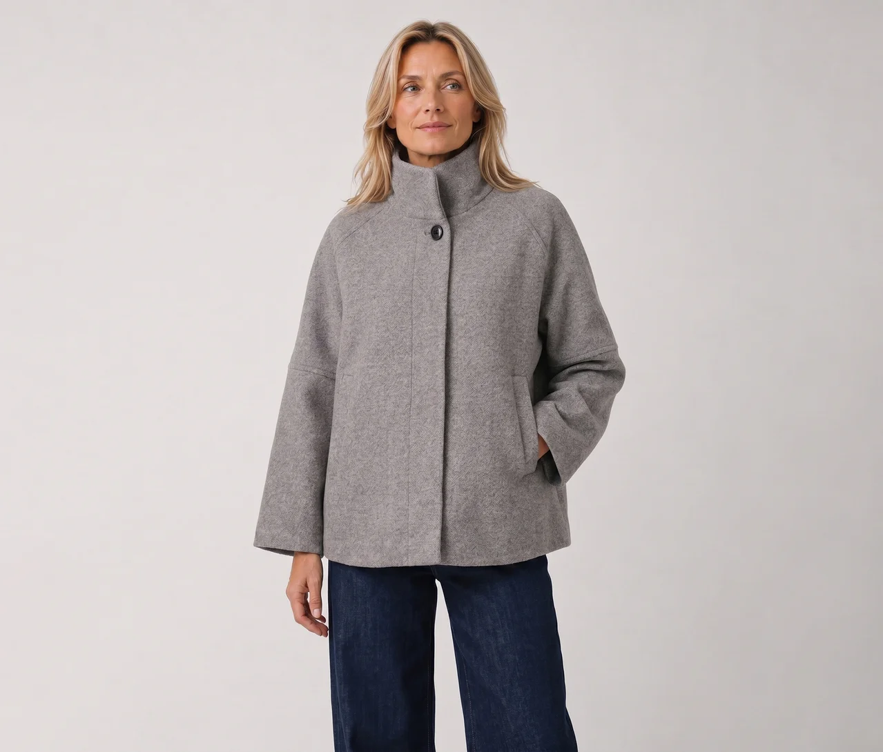 Eine blonde Frau steht in einer grauen, hochgeschlossenen Jacke mit weiten Ärmeln und dunklen Jeans, die rechte Hand in der Tasche.
