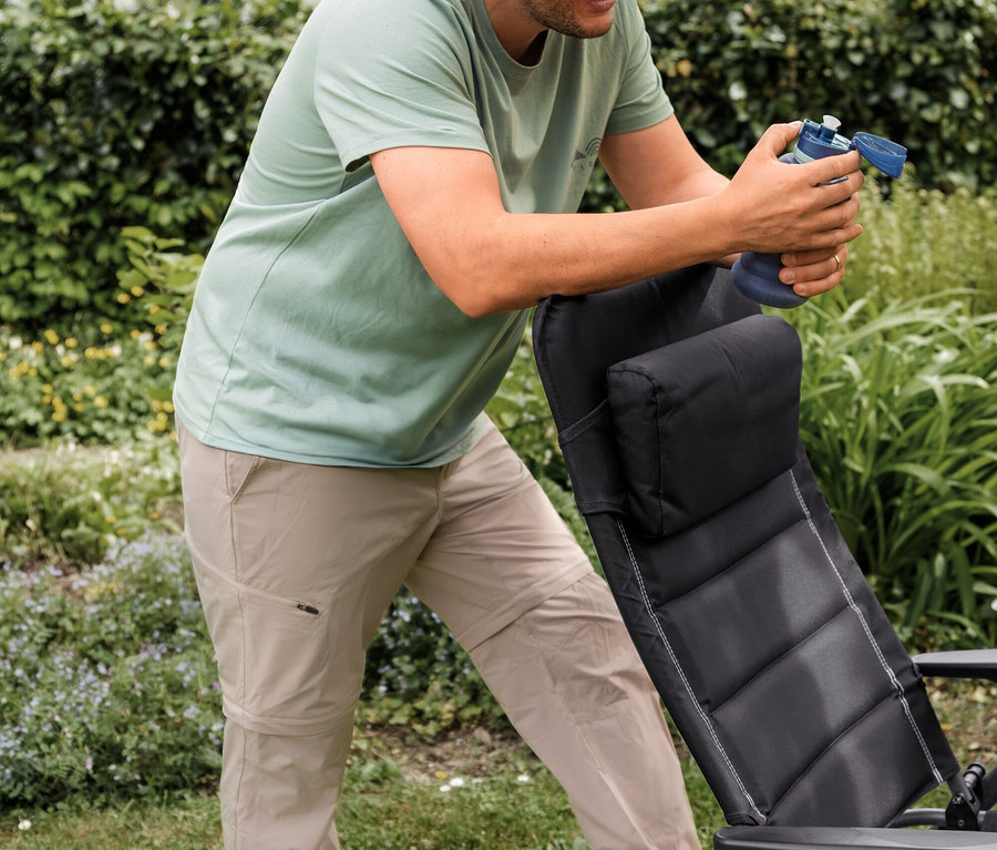 Lächelnder Mann in mintgrünem Outdoor-T-Shirt und beiger 2-in-1-Outdoor-Funktionshose, der sich auf einen schwarzen Gartenstuhl stützt und eine blaue Wasserflasche hält.