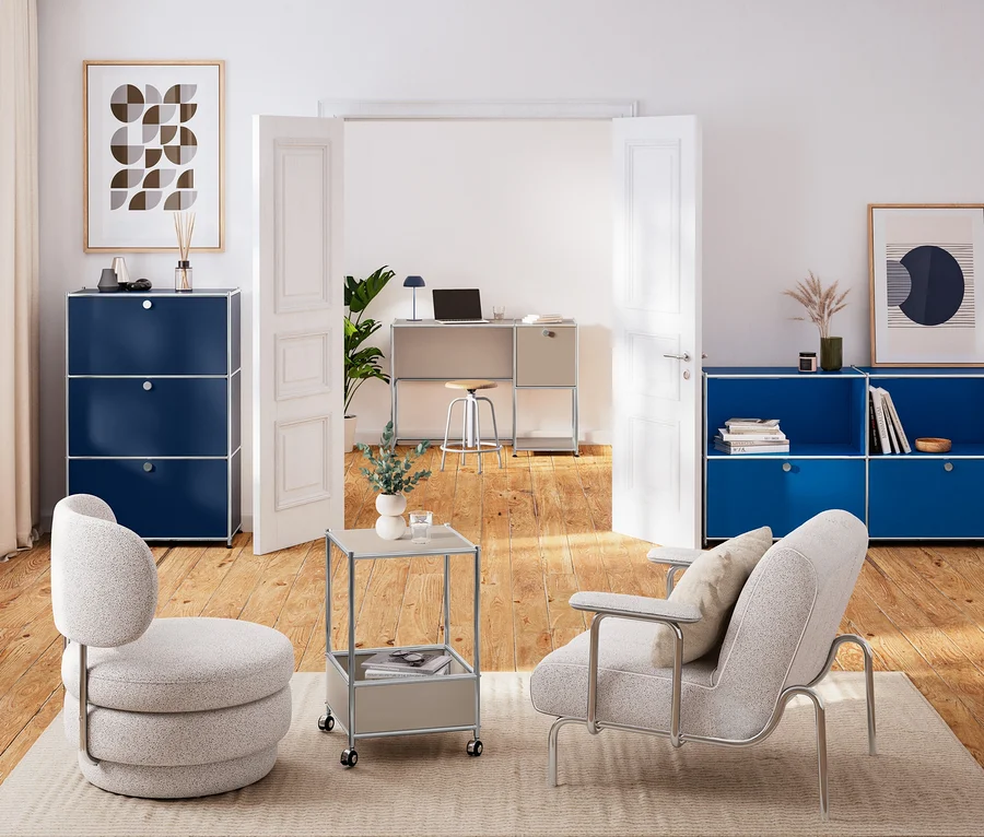 Zwei helle Sessel auf Teppich. Links dunkelblaue Kommode Metall »CN3«, rechts enzianblaues Sideboard Metall »CN3«. Dazwischen taupefarbener Couchtisch Metall »CN3«. Im Hintergrund taupefarbener Schreibtisch Metall »CN3« mit LED-Akku-Tischleuchte und Hocker.