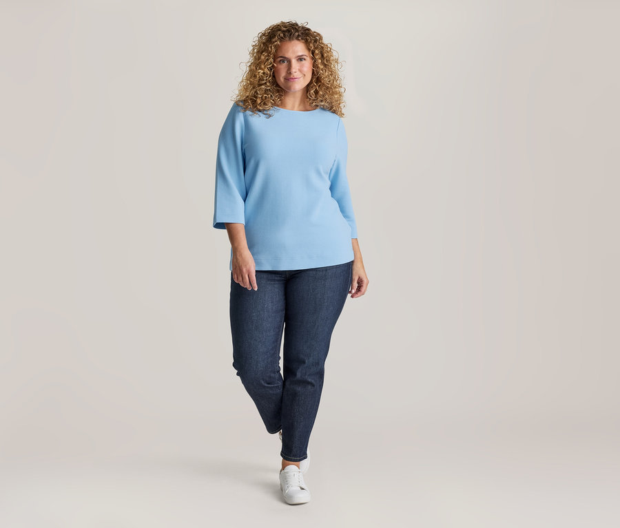 Frau mit lockigem Haar, hellblauem Oberteil mit 3/4-Ärmeln, dunkler Jeans und weißen Sneakern, lächelt in die Kamera.