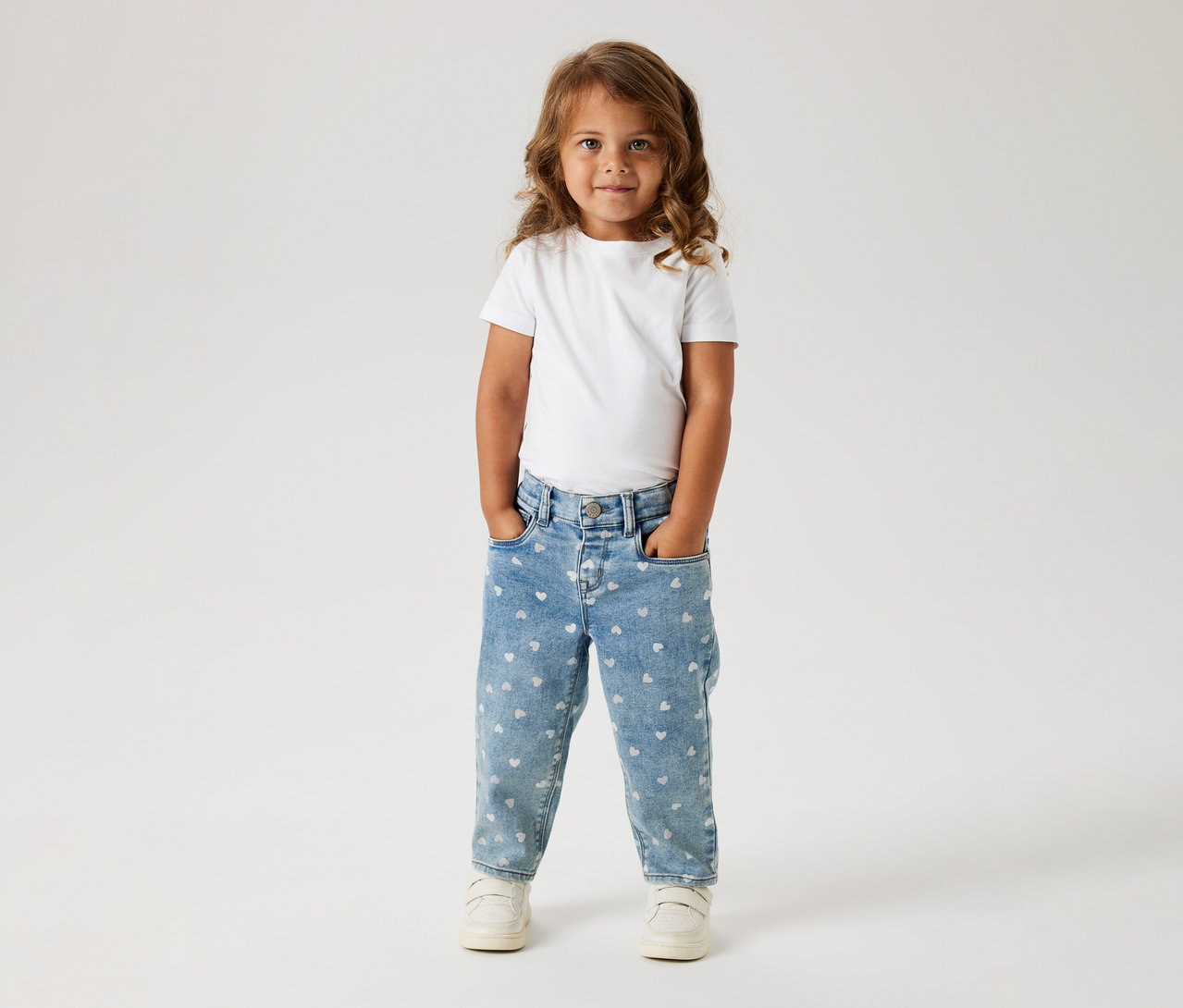 Ein kleines Kind mit langen, welligen braunen Haaren steht frontal, trägt ein weißes T-Shirt, hellblaue Jeans mit weißen Herzmustern und weiße Turnschuhe. Die Hände stecken in den Hosentaschen.