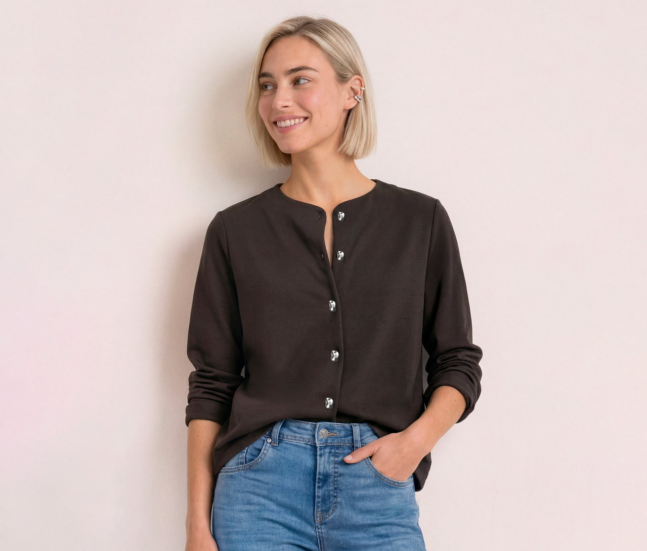 Junge Frau mit blondem Bob trägt einen dunkelbraunen Street One Cardigan mit silbernen Knöpfen und Jeans, lächelt zur Seite.