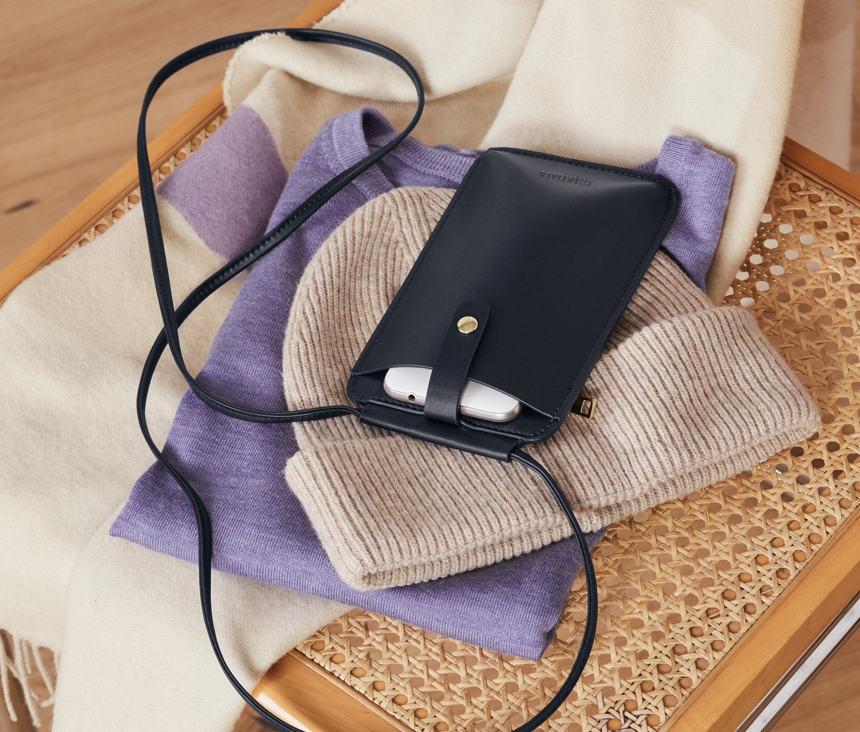 Auf einem Korbstuhl liegen eine marineblaue Handytasche, eine beige Mütze, ein lila Pullover und ein gestreifter Schal.