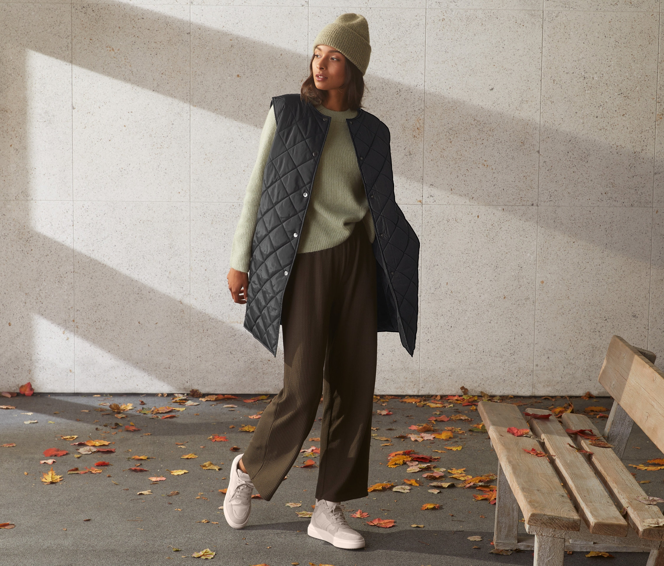 Frau in langer Steppweste, Pullover und Hose steht draußen neben einer Bank.