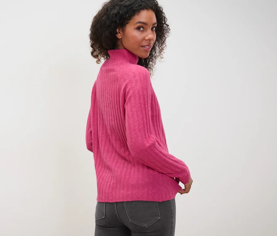Frau blickt über die Schulter und trägt einen gerippten Strickpullover mit Stehkragen und graue Jeans.