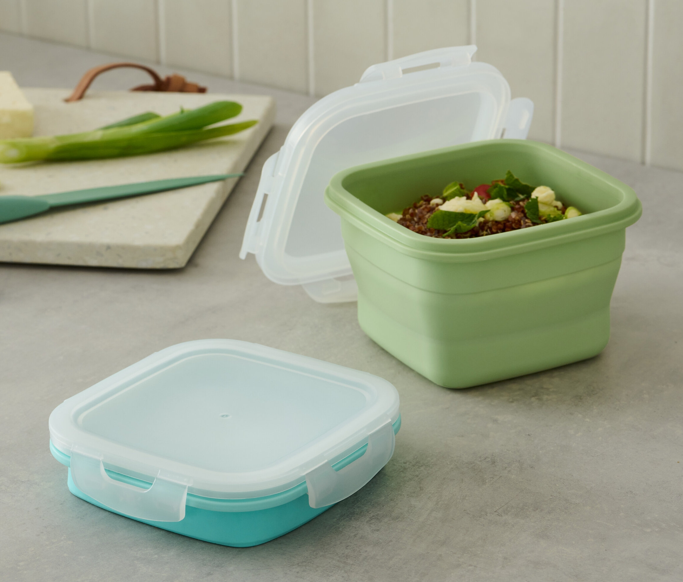 2 Frischhaltedosen stehen auf einem Tisch. Eine Dose ist grün und offen, die andere ist türkis und geschlossen.