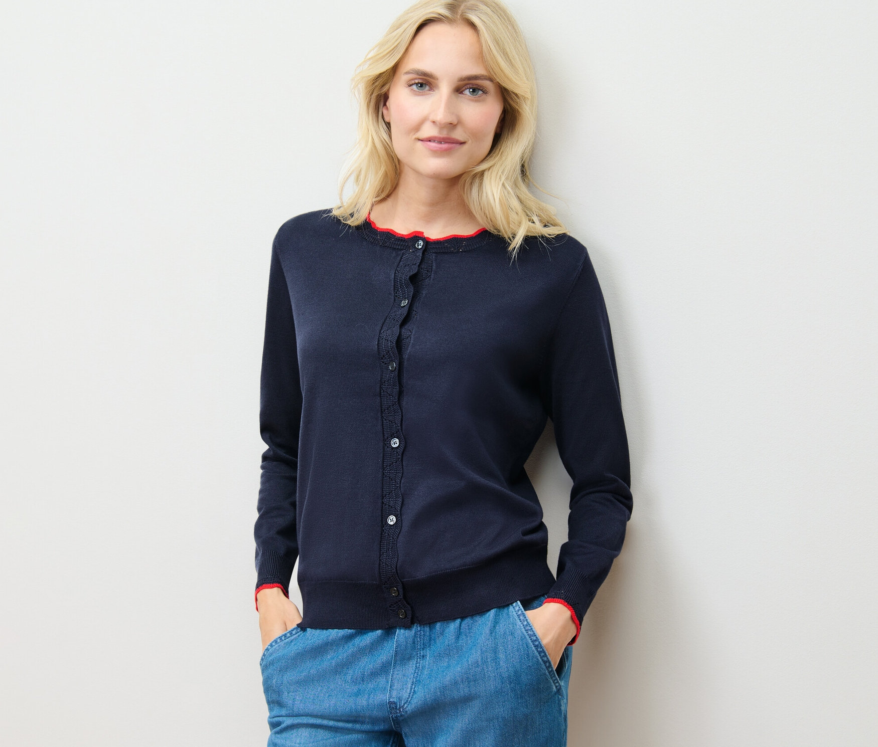 Eine Frau trägt einen Feinstrick-Cardigan mit Wellenkante und blaue Hosen und steht an einer Wand.