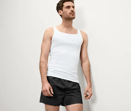Mann lehnt an Wand, trägt weißes Feinripp-Unterhemd und schwarze Boxershorts.