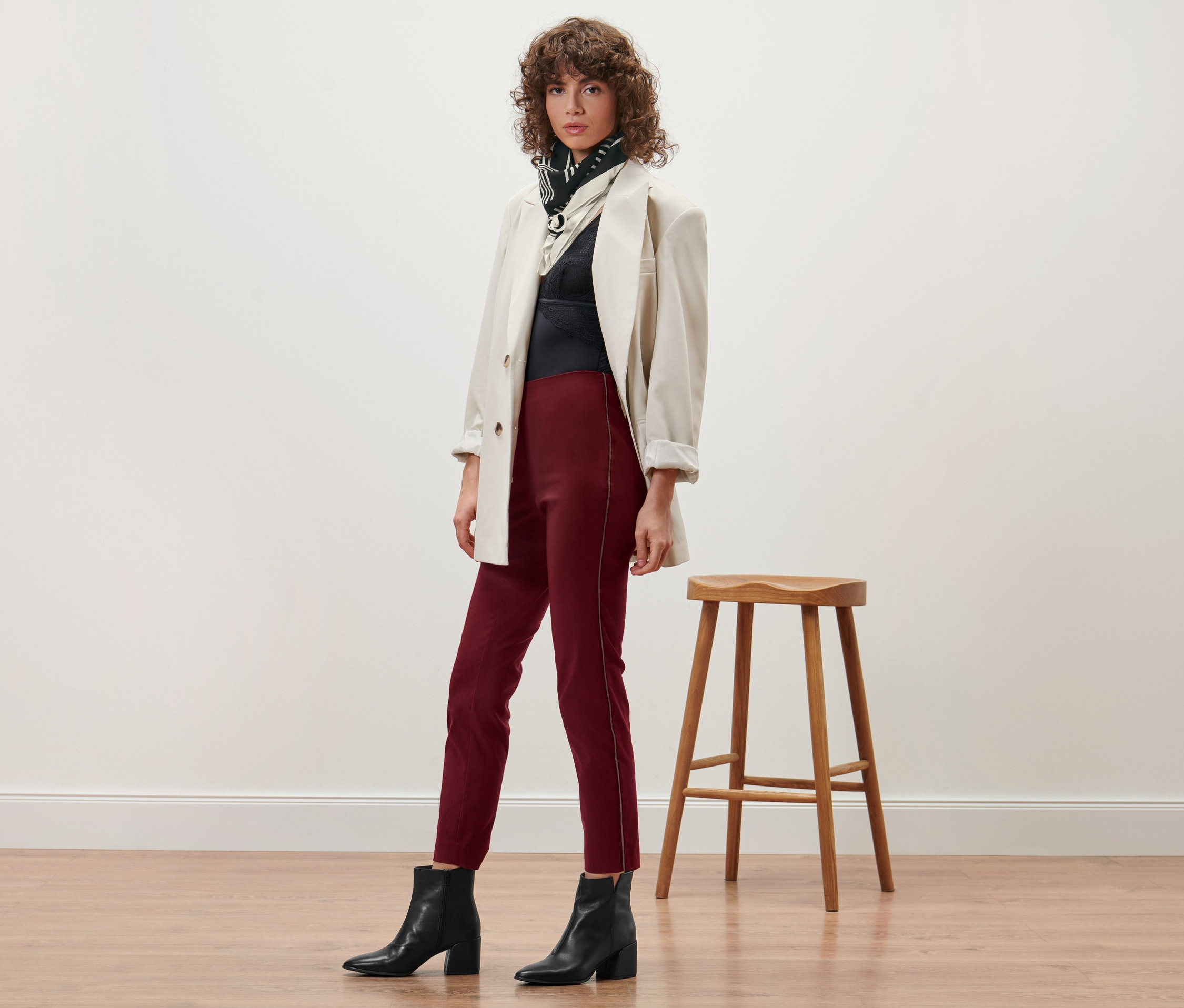 Frau posiert in bordeauxroten Comfort-Hosen, schwarzem Oberteil, beige-farbenem Blazer und schwarzen Stiefeln.
