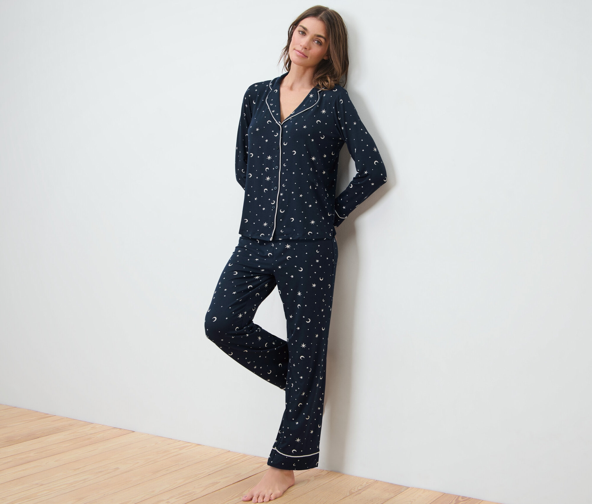 Eine Frau, die sich an eine weiße Wand lehnt, trägt einen dunkelblauen Pyjama mit Sternenhimmel.