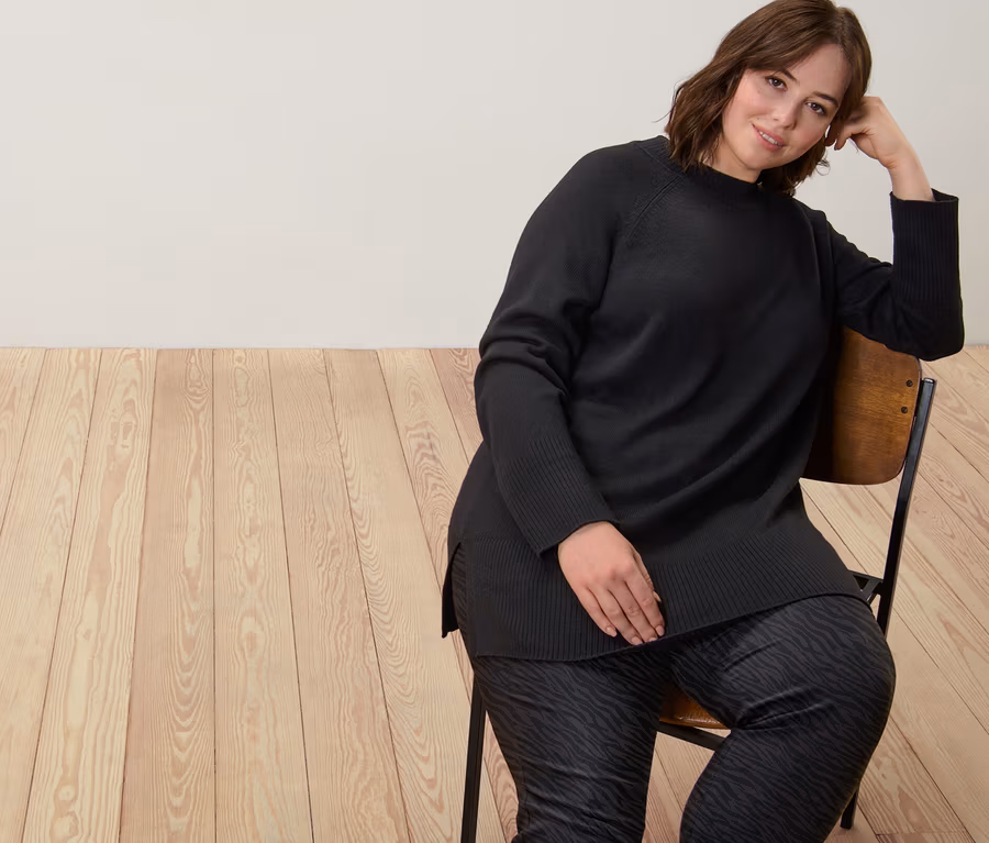 Frau sitzt auf einem Stuhl und trägt einen langen schwarzen Strickpullover und eine bedruckte Bengalin-Hose.