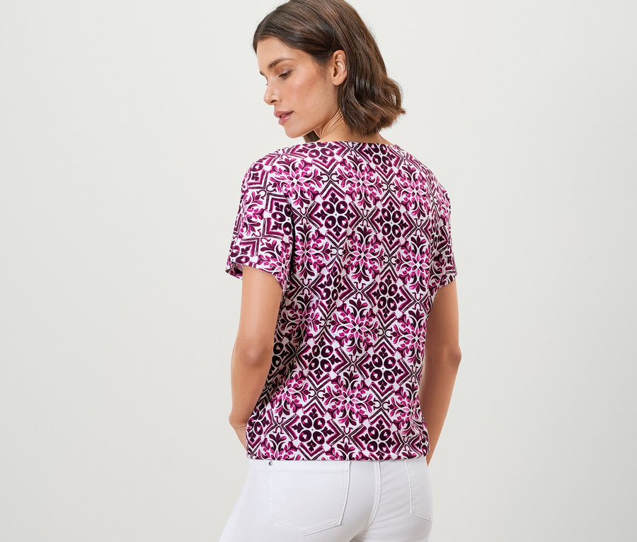 Frau posiert in einem bedruckten Blusenshirt in Berry-Farbe und weißen Hosen.