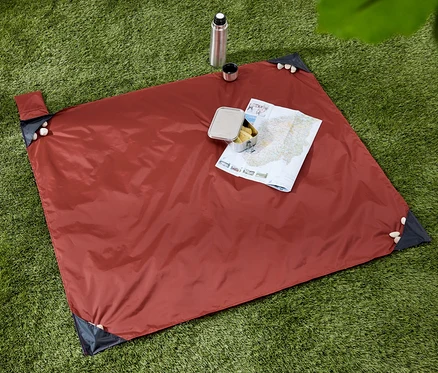 Auf grünem Gras liegt eine Mini-Picknickdecke. Auf der Decke liegen eine Landkarte und eine Brotbox. Eine Thermoskanne mit Becher steht daneben.