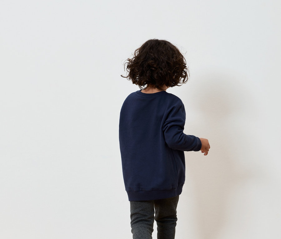 Junge mit braunen lockigen Haaren in marineblauem Sweatshirt und grauer Hose steht mit dem Rücken zu einer weißen Wand.