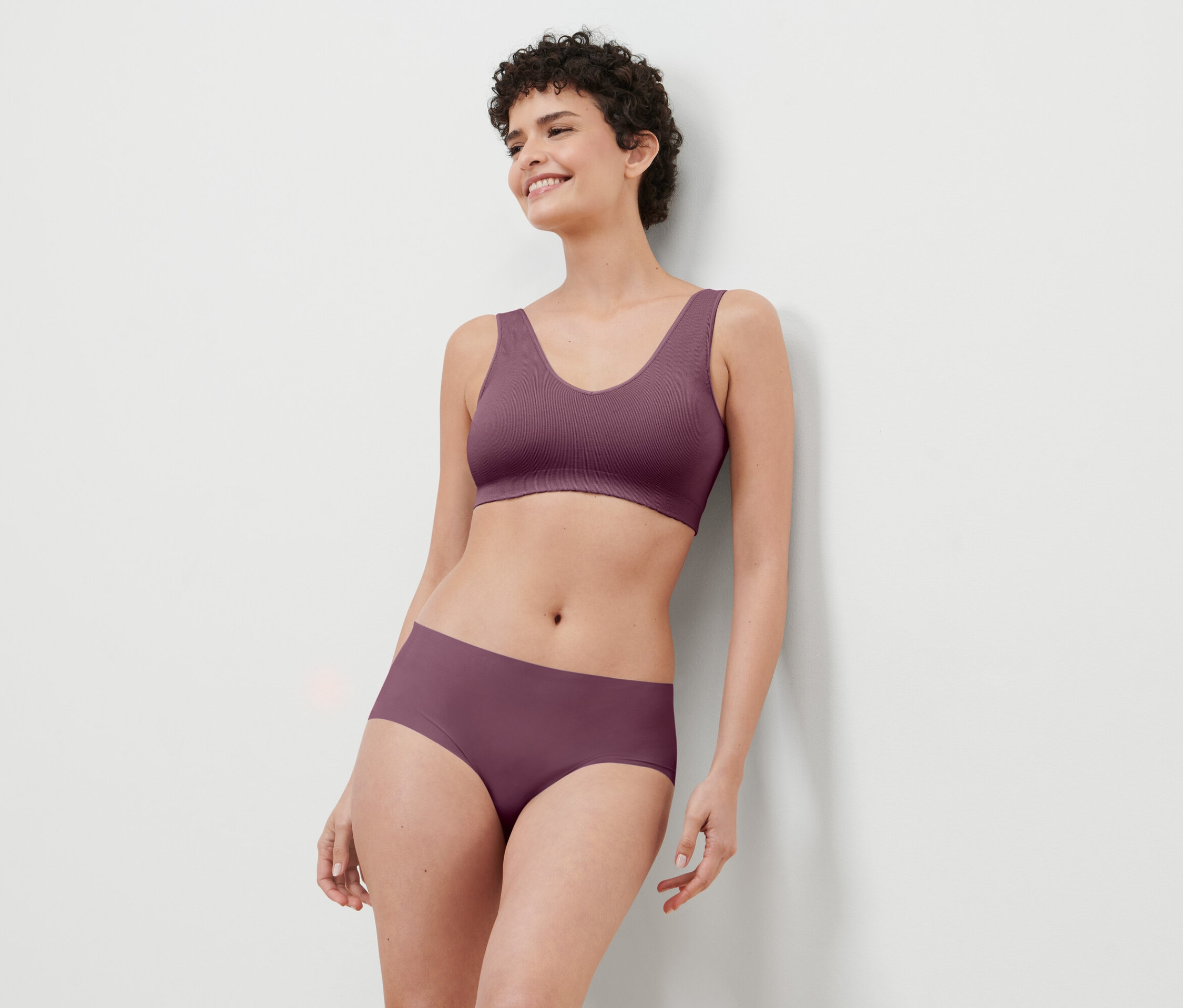 Frau mit kurzen Haaren, die ein bordeauxfarbenes Wäscheset trägt, lehnt an einer weißen Wand und lächelt.