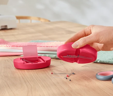 Eine Hand hält ein magnetisches Nadelkissen über einem geöffneten magnetischen Nadelkissen, daneben liegen eine Schere und ein Stoffstapel.