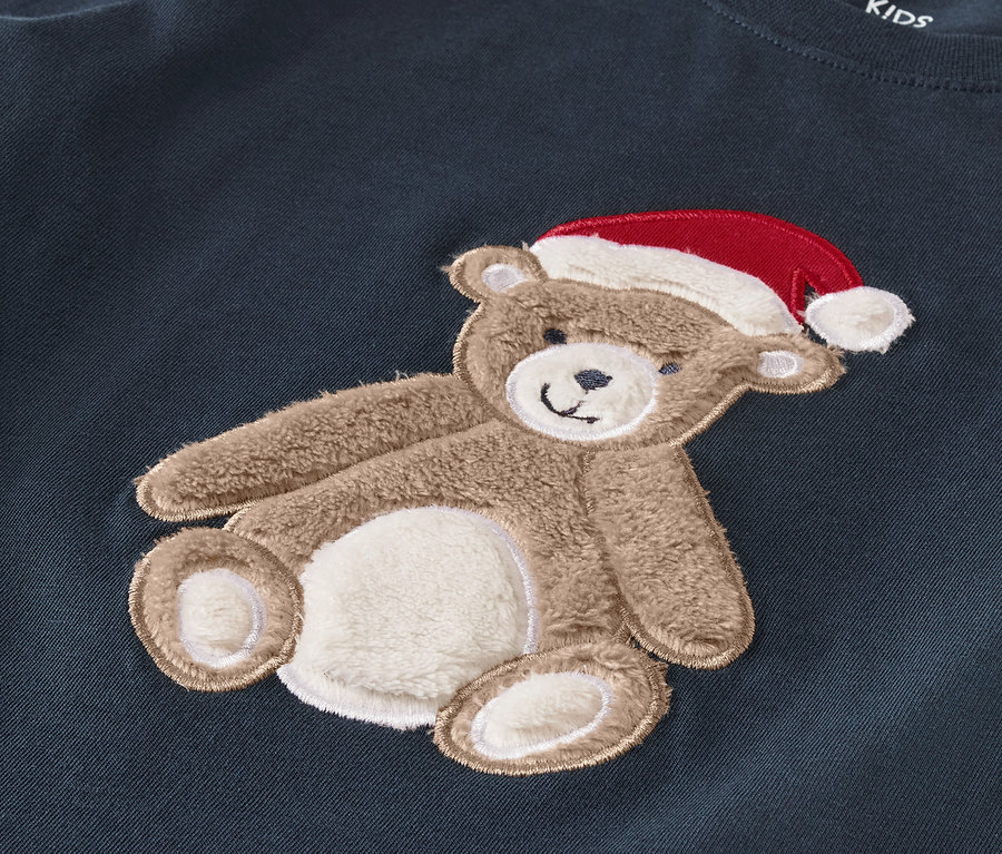 Stickerei eines Teddybären mit roter Mütze auf dunkelblauer Kleidung.